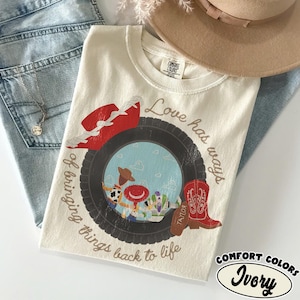 May include: An ivory t-shirt featuring a graphic of a black tire framing a scene with a cowboy doll and a space ranger toy. The design includes a red cowboy hat, a red boot, and the text Love has ways of bringing things back to life.