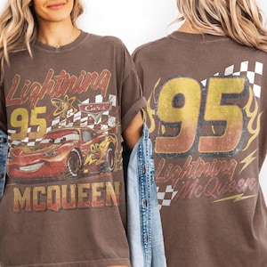 Może przedstawiać: Brązowa koszulka z Lightning McQueen z filmu Auta. Prz&oacute;d przedstawia numer 95, postać i szachownicę. Tył ma numer 95 i napis "Lightning McQueen".
