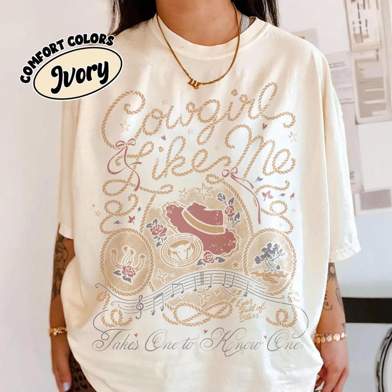 May include: An ivory t-shirt featuring a graphic design with the text "Cowgirl Like Me" and "Takes One to Know One" in a rope-style font. The print includes a cowboy hat, a horseshoe, a bull skull, musical notes, and small floral accents.