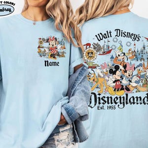 Peut inclure: T-shirt bleu clair avec Minnie Mouse et le mot "Name" sur le devant. Le dos du t-shirt pr&eacute;sente une illustration color&eacute;e de personnages Disney et le texte "Walt Disney's Disneyland Est. 1955".