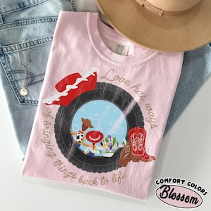 May include: A light pink t-shirt featuring a graphic of a black tire framing a blue sky with clouds and three cartoon characters. The design includes a red cowboy hat, a red boot, and the text Love has ways of bringing things back to life.