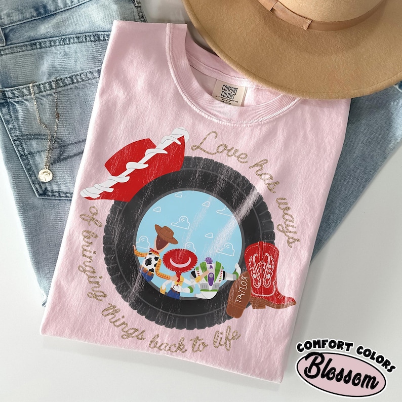 May include: A light pink t-shirt featuring a graphic of a black tire framing a blue sky with clouds and three cartoon characters. The design includes a red cowboy hat, a red boot, and the text Love has ways of bringing things back to life.