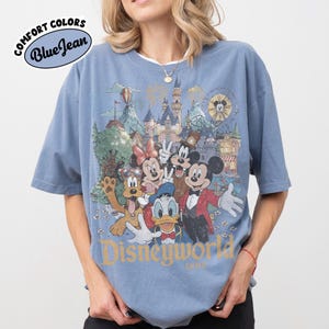 May include: Blue t-shirt with a graphic of Disney characters, including Mickey Mouse, Minnie Mouse, and Donald Duck, in front of a castle and amusement park. The shirt has the words "Disneyworld" and "Comfort Colors Blue Jean" printed on it.