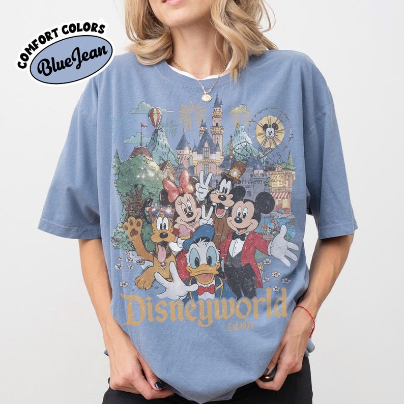 May include: Blue t-shirt with a graphic of Disney characters, including Mickey Mouse, Minnie Mouse, and Donald Duck, in front of a castle and amusement park. The shirt has the words "Disneyworld" and "Comfort Colors Blue Jean" printed on it.