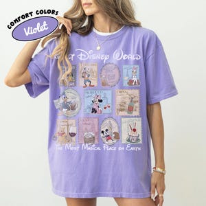 May include: Violet Comfort Colors t-shirt featuring a colorful graphic of Disney World stamps. The stamps showcase various Disney characters and international landmarks. Text includes "Walt Disney World" and "The Most Magical Place on Earth."