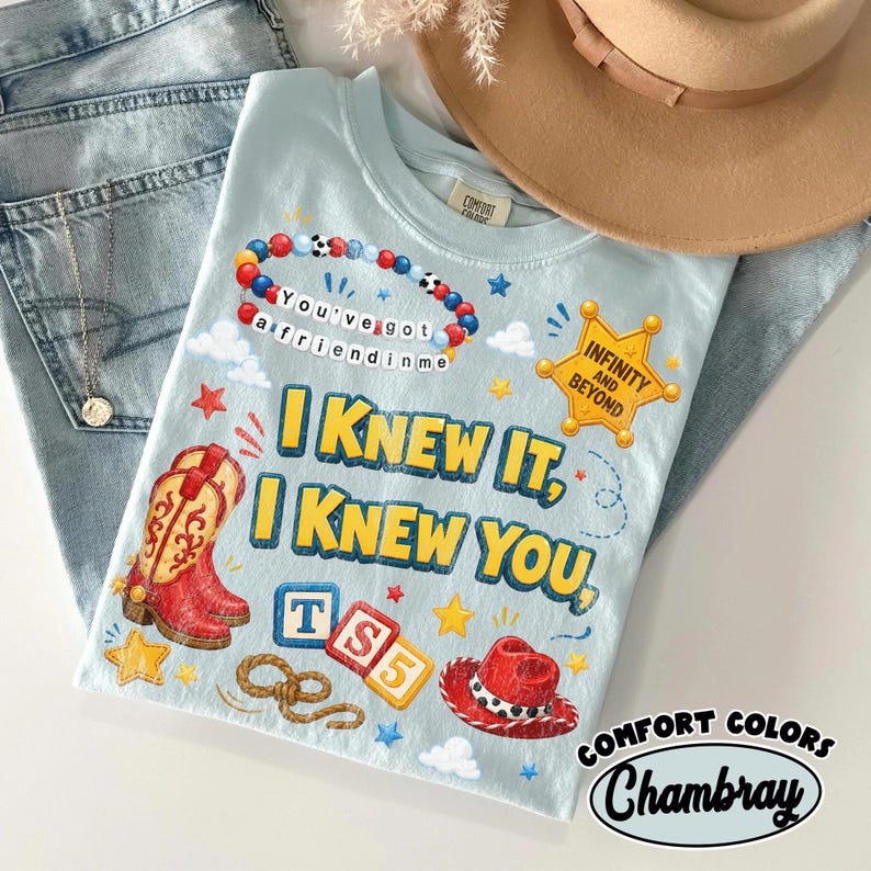 May include: A light blue t-shirt featuring a graphic print with a red cowboy boot, a red cowboy hat, a beaded bracelet with the text You've got a friend in me, a sheriff badge with Infinity and Beyond, and the text I KNEW IT, I KNEW YOU, TS5.