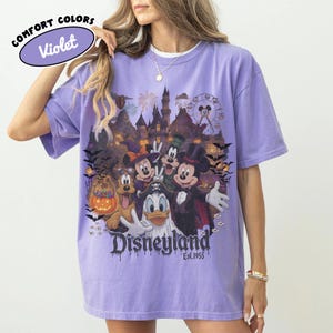May include: A violet short-sleeve t-shirt featuring a graphic print of Disney characters in Halloween costumes, including Mickey Mouse, Minnie Mouse, Goofy, Pluto, and Donald Duck in front of a castle, with the text Disneyland Est. 1955.