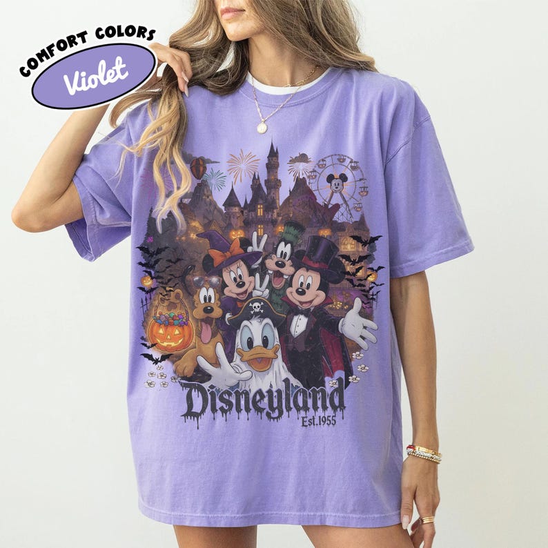 May include: A violet short-sleeve t-shirt featuring a graphic print of Disney characters in Halloween costumes, including Mickey Mouse, Minnie Mouse, Goofy, Pluto, and Donald Duck in front of a castle, with the text Disneyland Est. 1955.