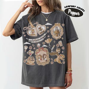 May include: Dark gray Comfort Colors t-shirt featuring Chip and Dale cartoon graphics in ornate frames, hearts, and clouds. The shirt includes the text "Dooney & Bourke" and "Comfort Colors Pepper".