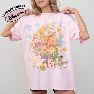 May include: A light pink t-shirt featuring a graphic print of various animated characters in a collage style. The design includes the letters TS, clouds, a rocket, a sheriff star, and a colorful ball. The text Comfort Colors Blossom is visible.