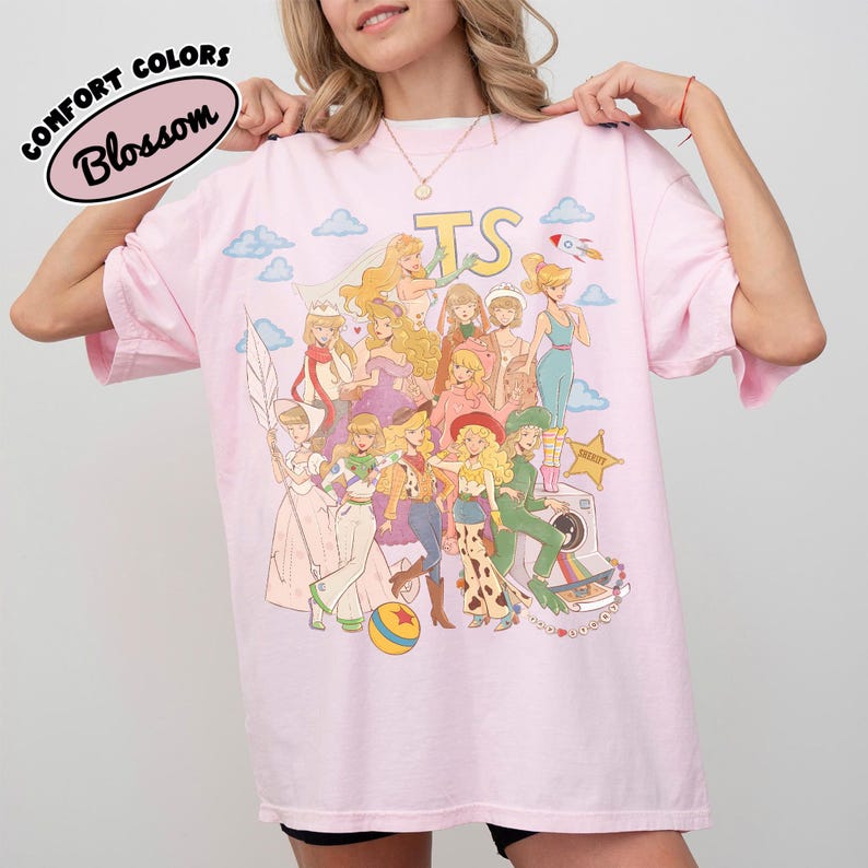 May include: A light pink t-shirt featuring a graphic print of various animated characters in a collage style. The design includes the letters TS, clouds, a rocket, a sheriff star, and a colorful ball. The text Comfort Colors Blossom is visible.