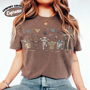 K&ouml;nnte beinhalten: Ein dunkelbraunes Kurzarm-T-Shirt mit einem Grafikdruck von Star Wars-Figuren wie Chewbacca, einem Stormtrooper, Grogu, R2-D2, BB-8 und Darth Vader, inklusive Luftballons und Lichtschwertern. Der Text Comfort Colors Espresso steht oben links.