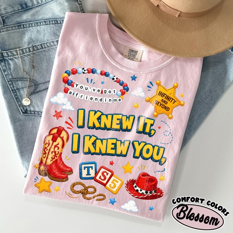 May include: A light pink t-shirt featuring a graphic print with a beaded bracelet reading You've got a friend in me, a yellow sheriff badge with Infinity and Beyond, red cowboy boots, a red cowboy hat, and blocks spelling TS5 with the text I KNEW IT, I KNEW YOU.
