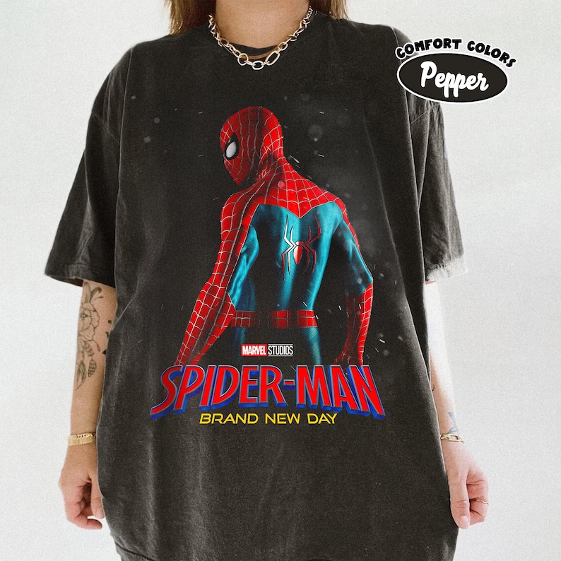 May include: Dark gray t-shirt featuring a graphic of Spider-Man in a red and blue suit, with the text "SPIDER-MAN BRAND NEW DAY" in red and blue. The shirt also has the text "COMFORT COLORS Pepper" in a white oval.