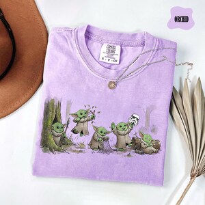 May include: A light purple short-sleeve t-shirt featuring a graphic print of five small green creatures in robes interacting with a forest setting and a white helmet. The shirt has a Comfort Colors brand tag at the neck and the text ORCHID.