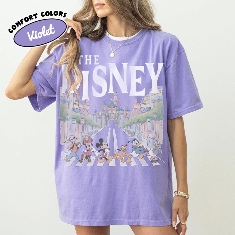 Może przedstawiać: Fioletowa koszulka z białym napisem "THE DISNEY". Koszulka przedstawia grafikę postaci Disneya przechodzących przez ulicę przed zamkiem. Słowa "COMFORT COLORS" i "Violet" znajdują się w fioletowym owalu.