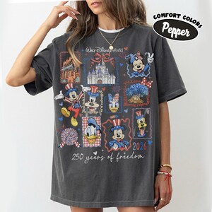 May include: Dark gray t-shirt featuring a colorful graphic of Disney characters and landmarks. The design includes Mickey Mouse, Minnie Mouse, Donald Duck, and Goofy, with text that reads "Walt Disney World" and "250 years of freedom" and the year "2026".