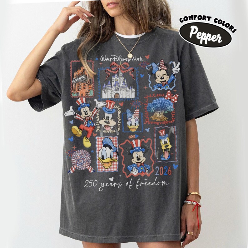 May include: Dark gray t-shirt featuring a colorful graphic of Disney characters and landmarks. The design includes Mickey Mouse, Minnie Mouse, Donald Duck, and Goofy, with text that reads "Walt Disney World" and "250 years of freedom" and the year "2026".