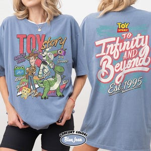 Pode incluir: Uma t-shirt azul com gr&aacute;ficos de Toy Story. A frente mostra Woody, Buzz Lightyear, Rex e Smarty Pants com texto incluindo Toy Story, Since 1995, Hi, Lilypad, Snappy e Woody. As costas apresentam o texto To Infinity and Beyond Est. 1995.