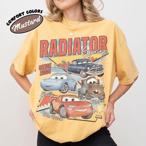 May include: Mustard-colored t-shirt featuring a graphic of characters from the movie Cars, including Lightning McQueen, Sally, Mater, and Doc Hudson. The text "RADIATOR SPRINGS" and "SINCE 2006" are also displayed. The shirt is a Comfort Colors brand.