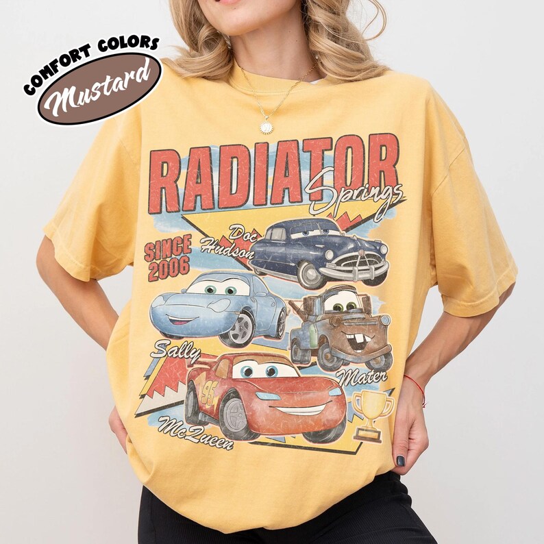 May include: Mustard-colored t-shirt featuring a graphic of characters from the movie Cars, including Lightning McQueen, Sally, Mater, and Doc Hudson. The text "RADIATOR SPRINGS" and "SINCE 2006" are also displayed. The shirt is a Comfort Colors brand.