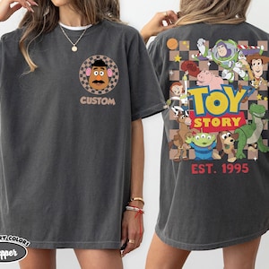 Puede incluir: Camiseta gris oscuro con un dise&ntilde;o de Mr. Potato Head y la palabra "CUSTOM" en la parte delantera. La parte trasera muestra un gr&aacute;fico de Toy Story con varios personajes y el texto "TOY STORY EST. 1995".