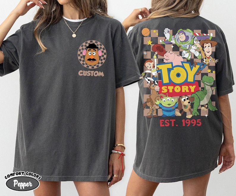Puede incluir: Camiseta gris oscuro con un dise&ntilde;o de Mr. Potato Head y la palabra "CUSTOM" en la parte delantera. La parte trasera muestra un gr&aacute;fico de Toy Story con varios personajes y el texto "TOY STORY EST. 1995".