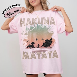 May include: A pale pink t-shirt with the words "HAKUNA MATATA" in a tan font. The shirt features a graphic of characters from The Lion King, including Simba, Timon, Pumbaa, and a bird. The words "Comfort Colors" and "Blossom" are also printed on the shirt.