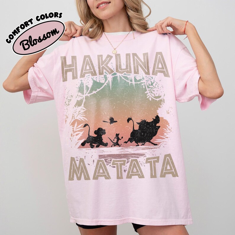 May include: A pale pink t-shirt with the words "HAKUNA MATATA" in a tan font. The shirt features a graphic of characters from The Lion King, including Simba, Timon, Pumbaa, and a bird. The words "Comfort Colors" and "Blossom" are also printed on the shirt.