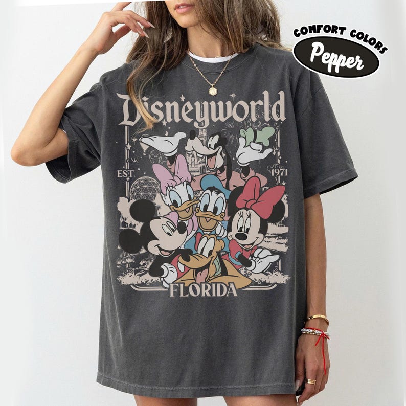 May include: Dark gray t-shirt featuring a graphic of Disney characters including Mickey, Minnie, Donald, and Daisy Duck. The shirt has the text "Disneyworld Florida" and "EST. 1971". The shirt is labeled "Comfort Colors Pepper".