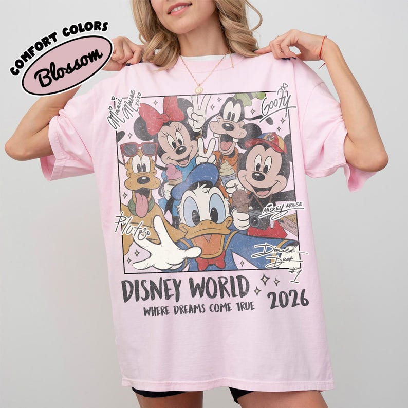 May include: A light pink t-shirt featuring a graphic of Disney characters including Mickey Mouse, Minnie Mouse, Donald Duck, and Goofy. The shirt has the words "DISNEY WORLD" and "2026" printed on it. The shirt is made of a soft material.