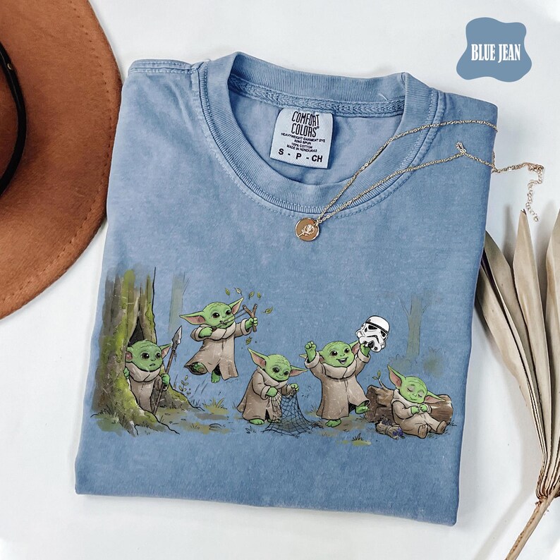 May include: A blue jean colored t-shirt featuring a graphic print of five small green creatures in robes. One creature holds a slingshot, one holds a net, one holds a stormtrooper helmet, and one sits by a log. The shirt has a Comfort Colors label.