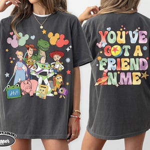 May include: A charcoal grey t-shirt featuring a front graphic of various animated toy characters and a green sign that says Hi. The back of the shirt displays the colourful text You've Got A Friend In Me with star, heart, and ball graphic accents.