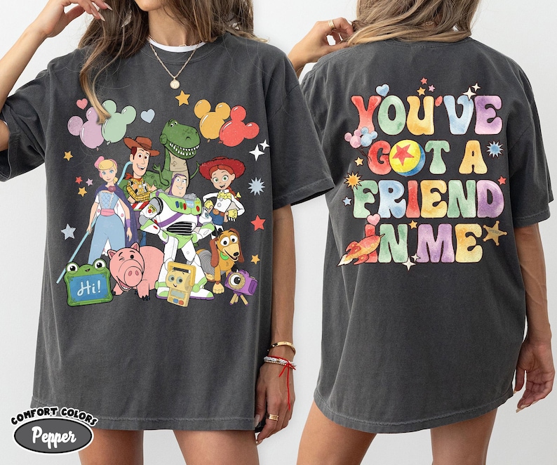 May include: A charcoal grey t-shirt featuring a front graphic of various animated toy characters and a green sign that says Hi. The back of the shirt displays the colourful text You've Got A Friend In Me with star, heart, and ball graphic accents.