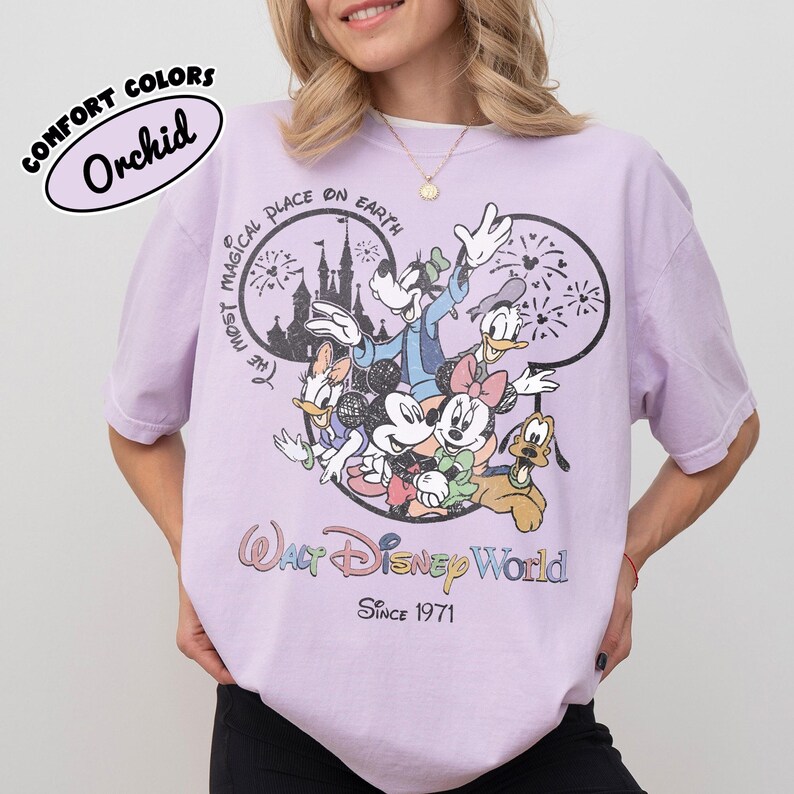 May include: Orchid-colored t-shirt featuring a graphic of Disney characters including Mickey, Minnie, Donald, Daisy, Goofy, and Pluto. The shirt has the text "Walt Disney World" and "Since 1971". The words "Comfort Colors" and "Orchid" are in a bubble on the top left.