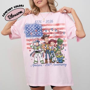 Może przedstawiać: R&oacute;żowa koszulka z grafiką amerykańskiej flagi i postaciami z kresk&oacute;wek. Koszulka ma napis "1776-2026" i "America - 250th Anniversary". Koszulka ma r&oacute;wnież napis "Comfort Colors Blossom".