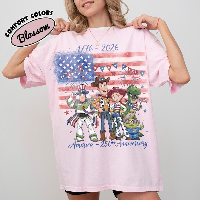 Może przedstawiać: R&oacute;żowa koszulka z grafiką amerykańskiej flagi i postaciami z kresk&oacute;wek. Koszulka ma napis "1776-2026" i "America - 250th Anniversary". Koszulka ma r&oacute;wnież napis "Comfort Colors Blossom".