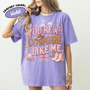 Puede incluir: Una camiseta violeta de manga corta con el texto "YOU'RE A COWGIRL LIKE ME" en letras rosas y marrones. El gr&aacute;fico incluye un sombrero de vaquero marr&oacute;n, una herradura, una bola de discoteca, un lazo rosa y botas con el texto "TOY STORY".