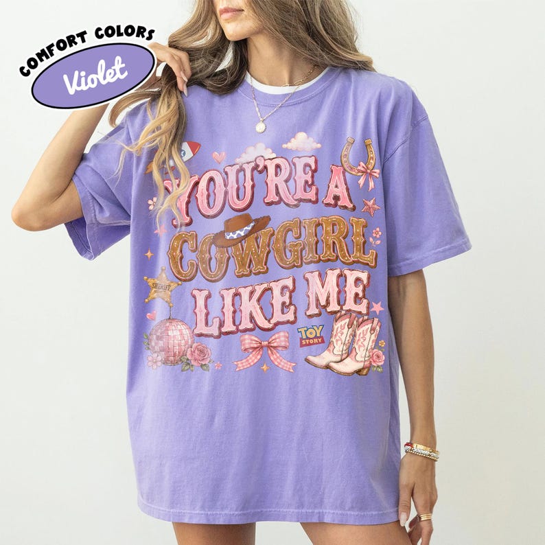 Puede incluir: Una camiseta violeta de manga corta con el texto "YOU'RE A COWGIRL LIKE ME" en letras rosas y marrones. El gr&aacute;fico incluye un sombrero de vaquero marr&oacute;n, una herradura, una bola de discoteca, un lazo rosa y botas con el texto "TOY STORY".