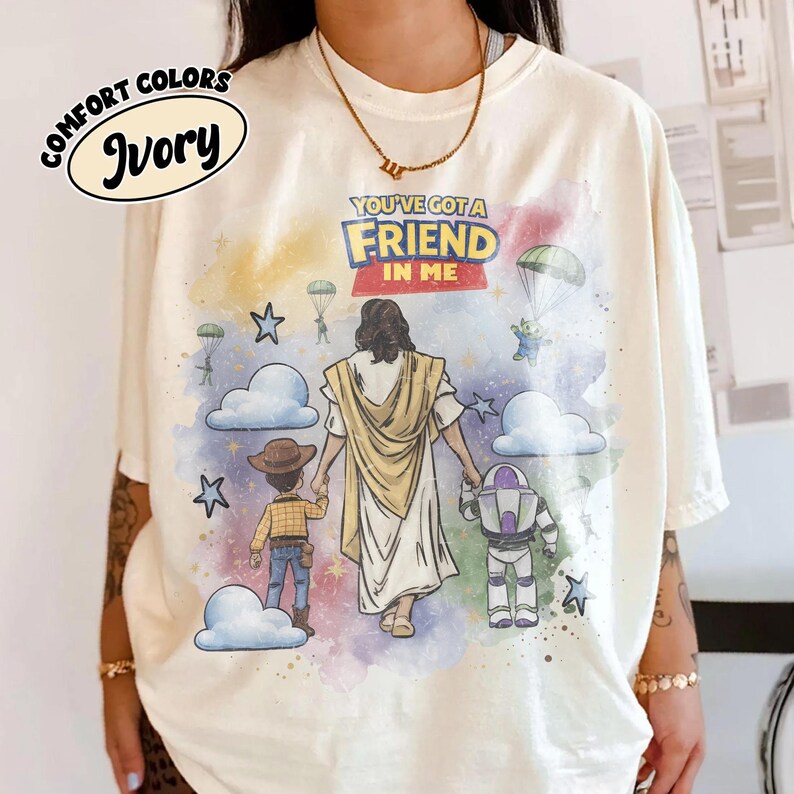 May include: Ivory Comfort Colors t-shirt featuring a colorful graphic with the text "YOU'VE GOT A FRIEND IN ME." The design includes illustrations of Jesus, Woody, and Buzz Lightyear, with clouds, stars, and parachutes.