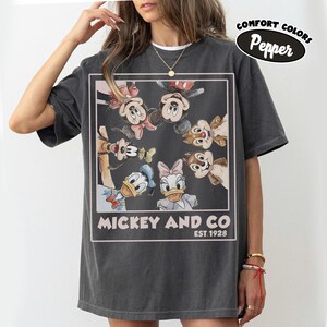Puede incluir: Camiseta gris oscuro con un gr&aacute;fico de Mickey Mouse, Minnie Mouse, Pato Donald, Daisy, Goofy y Chip y Chop. El gr&aacute;fico est&aacute; enmarcado con el texto "Mickey and Co. Est. 1928". La camiseta tiene una etiqueta Comfort Colors Pepper.