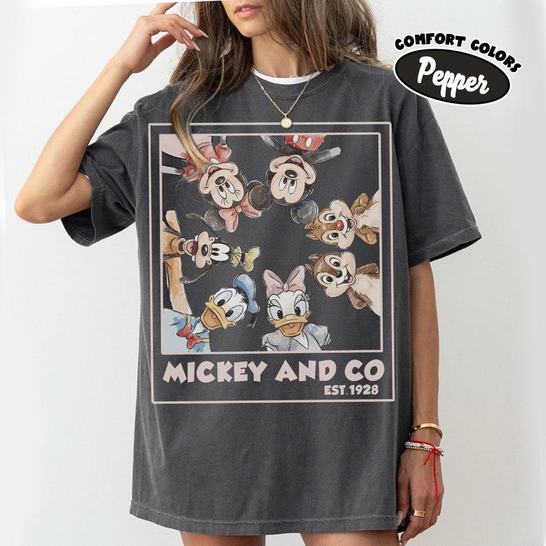 Puede incluir: Camiseta gris oscuro con un gr&aacute;fico de Mickey Mouse, Minnie Mouse, Pato Donald, Daisy, Goofy y Chip y Chop. El gr&aacute;fico est&aacute; enmarcado con el texto "Mickey and Co. Est. 1928". La camiseta tiene una etiqueta Comfort Colors Pepper.