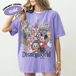 May include: Violet Comfort Colors t-shirt featuring a Disneyworld graphic with Mickey Mouse, Minnie Mouse, Donald Duck, Goofy, and other characters. The graphic includes a castle, Ferris wheel, and the text "Disneyworld Est.1971".