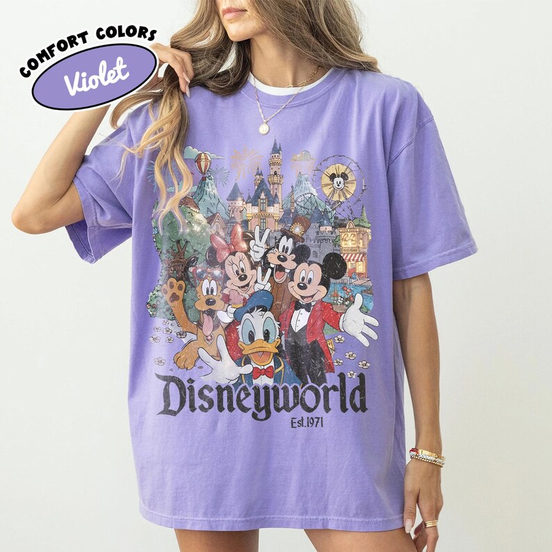May include: Violet Comfort Colors t-shirt featuring a Disneyworld graphic with Mickey Mouse, Minnie Mouse, Donald Duck, Goofy, and other characters. The graphic includes a castle, Ferris wheel, and the text "Disneyworld Est.1971".