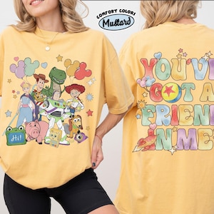 May include: A mustard yellow t-shirt featuring a front graphic of Toy Story characters including Woody, Buzz Lightyear, and Jessie with balloons. The back graphic displays the text "YOU'VE GOT A FRIEND IN ME" in colourful bubble letters with a rocket.