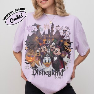 May include: A light purple t-shirt featuring a graphic of cartoon characters in Halloween costumes in front of a castle and Ferris wheel. The text reads COMFORT COLORS Orchid, Disneyland, and Est. 1955. The design includes fireworks and bats.