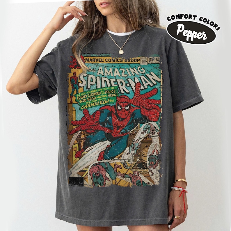 May include: A charcoal gray short-sleeve t-shirt featuring a vintage Marvel Comics The Amazing Spider-Man graphic print on the front. The design displays Spider-Man in a dynamic pose with comic book text and a distressed, weathered aesthetic.