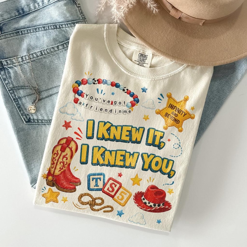 May include: A cream-colored t-shirt featuring a graphic print with the text "I KNEW IT, I KNEW YOU," a beaded bracelet with "YOU'VE GOT A FRIEND IN ME," a yellow sheriff star with "INFINITY AND BEYOND," cowboy boots, blocks, and a hat.
