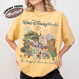May include: Mustard-colored t-shirt featuring a graphic of Walt Disney World with fireworks, a train, and various Toy Story characters. The text "Walt Disney World" and "The Happiest Place On Earth" are also visible.