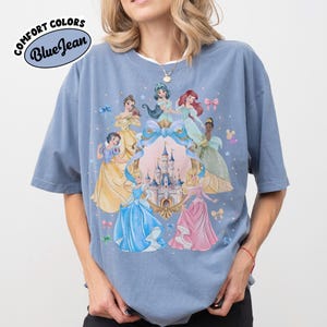 May include: Blue t-shirt with a graphic of Disney princesses surrounding a castle. The shirt has the words "Comfort Colors" and "Blue Jean" printed on it. The princesses are in various gowns, with a light blue background.